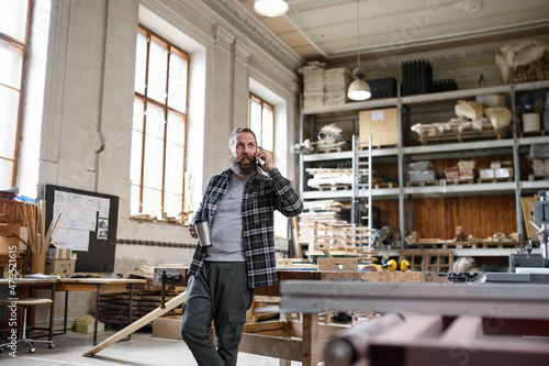 Happy mature male carpenter making phone call during coffee break indoors in carpentery workshop.