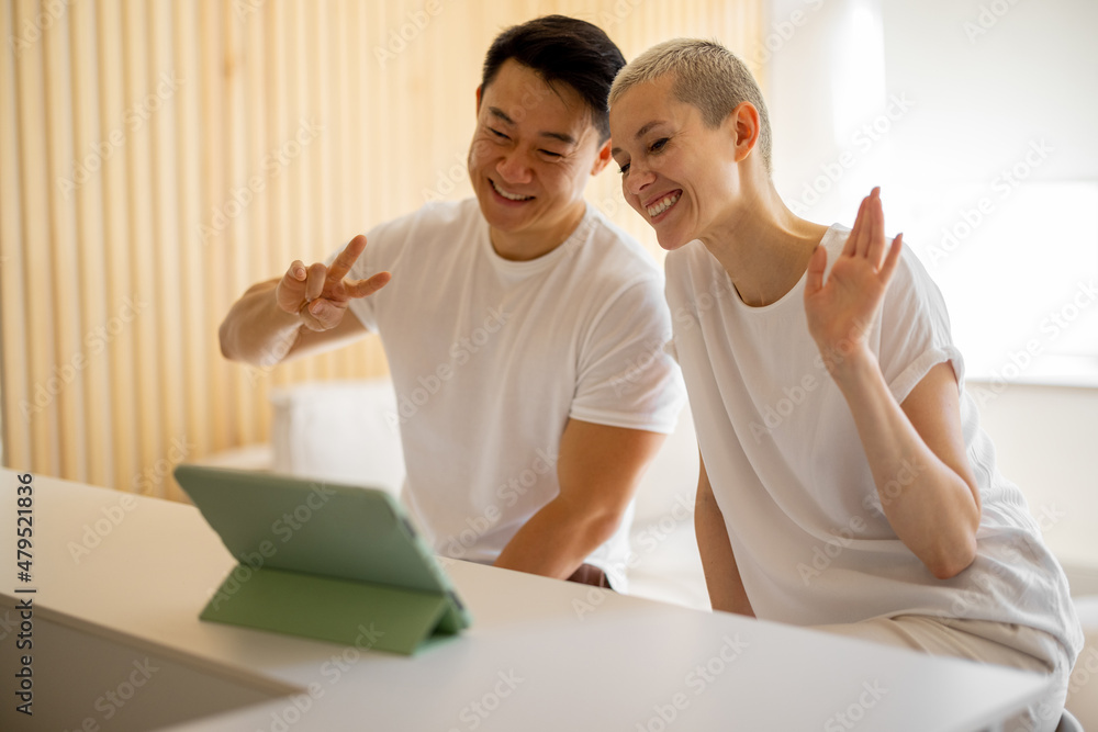 Smiling multiracial couple having video call on digital tablet at home. Concept of domestic lifestyle. Idea of online communication. Asian man showing victory gesture. European girl waving hand Smiling multiracial couple having video call on digital tablet at home. Concept of domestic lifestyle. Idea of online communication. Asian man showing victory gesture. European girl waving hand