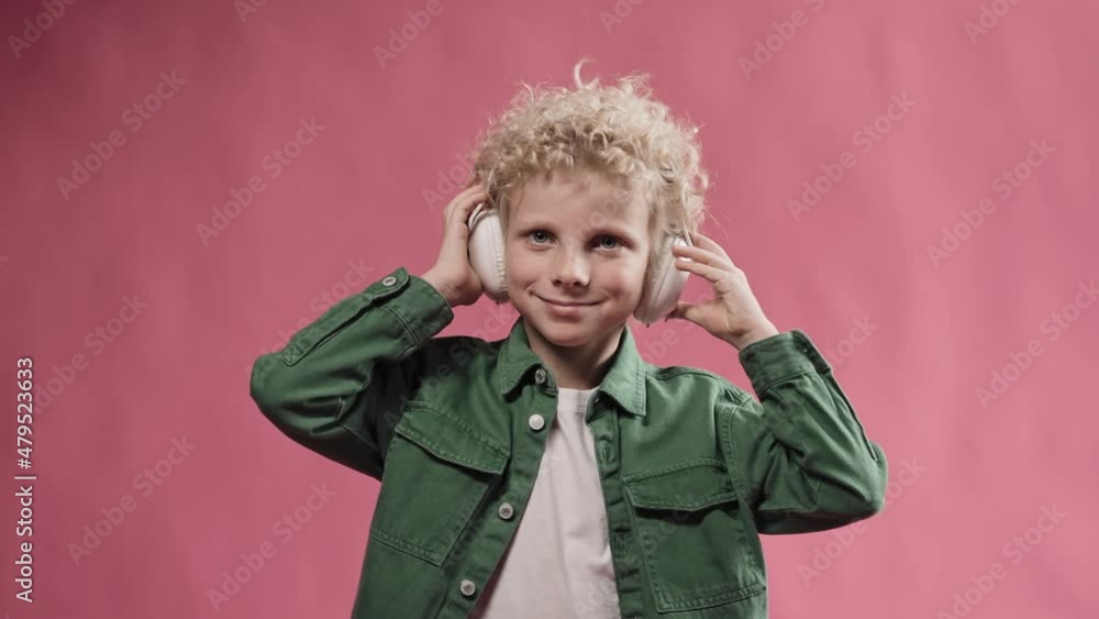 Little blonde boy wearing big headphones and nodding his head smiling. Cute kid smiling standing in the studio with pink background. High quality 4k footage Little blonde boy wearing big headphones and nodding his head smiling. Cute kid smiling standing in the studio with pink background. High quality 4k footage