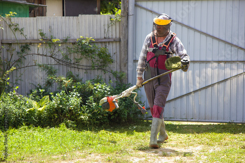 Russia, Vladivostok, August 22, 2018: Aged man in overalls with a lawn mower mows the lawn. The gardener cares for the garden. photo