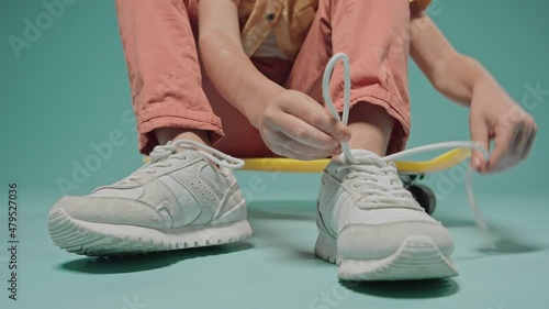Boy tying shoelaces on sneakers sitting on a skateboard on blue background. Cute kid sitting in the studio with blue background. High quality 4k footage