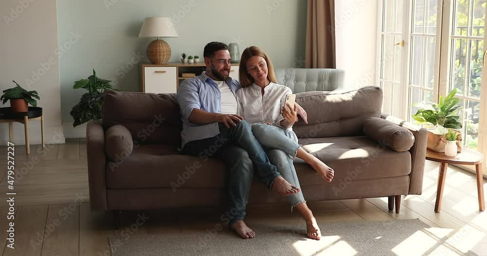 Happy couple in love relax on comfy couch at living room look on phone screen use app to shoot funny selfie video clip. Smiling young husband wife hug talk having fun with online applications on cell