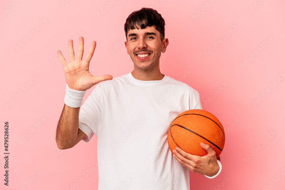 Young mixed race man playing basketball isolated on pink background smiling cheerful showing number five with fingers.