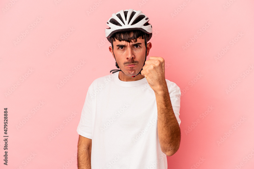 Young mixed race man wearing a helmet bike isolated on pink background showing fist to camera, aggressive facial expression. Young mixed race man wearing a helmet bike isolated on pink background showing fist to camera, aggressive facial expression.