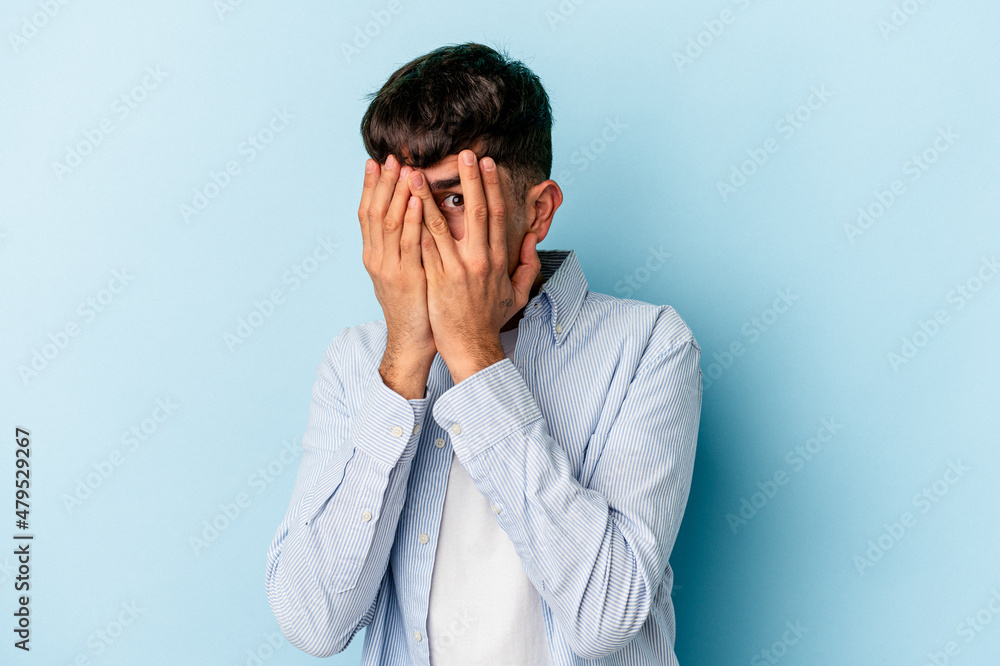 Young mixed race man isolated on blue background blink at the camera through fingers, embarrassed covering face. Young mixed race man isolated on blue background blink at the camera through fingers, embarrassed covering face.