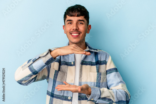 Young mixed race man isolated on blue background holding something with both hands, product presentation.