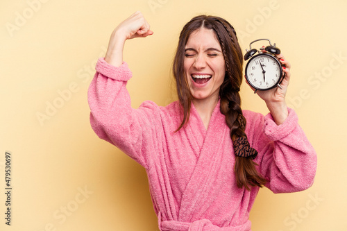 Young caucasian woman wearing a bathrobe holding a alarm clock isolated on yellow background raising fist after a victory, winner concept. © Asier