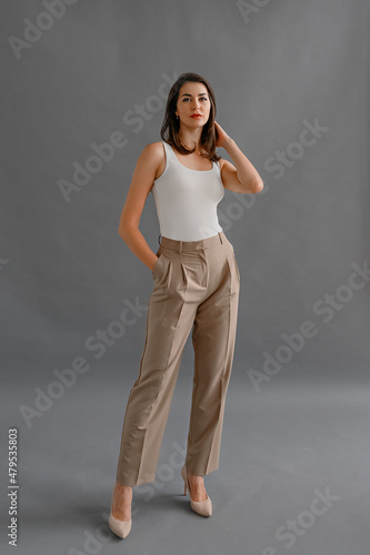 Full length portrait of happy businesswoman looking confident at camera, dressed in white t shirt with short sleeves and beige pants, posing in studio over gray background