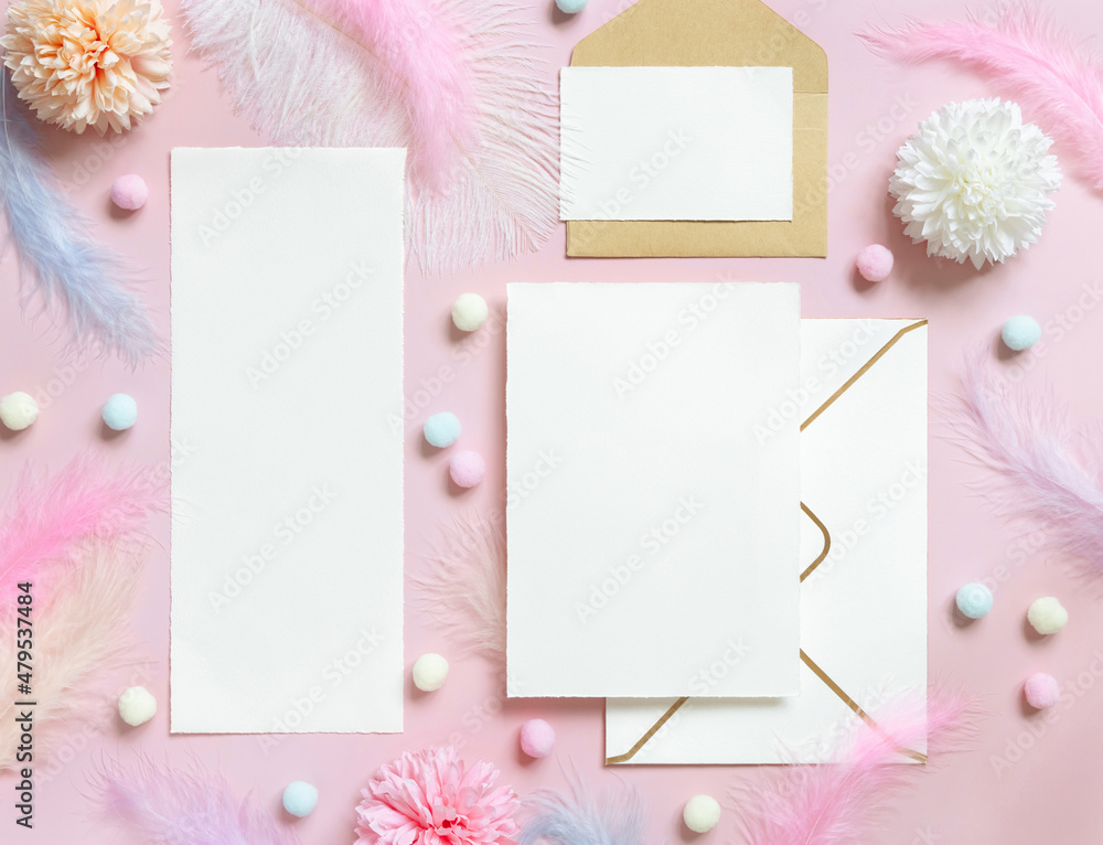 Blank cards and envelope near pastel flowers, pom-poms and feathers near ring in a gift box on pink
