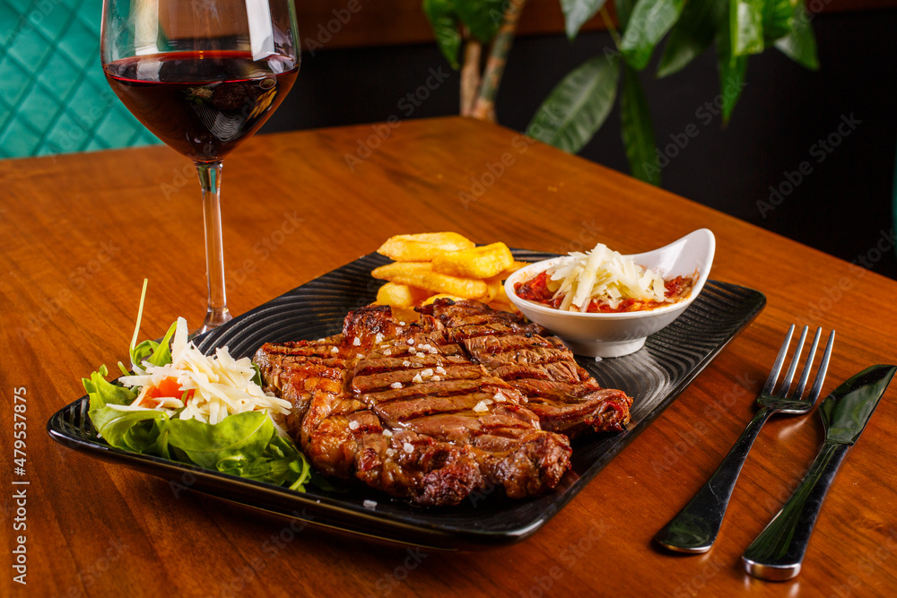 Bistecca di carne Ribeye di Scottona cotta al sangue con contorno di rucola, pomodorini, scaglie di formaggio, patate fritte e parmigiana di melanzane accompagnata da un calice di vino rosso Bistecca di carne Ribeye di Scottona cotta al sangue con contorno di rucola, pomodorini, scaglie di formaggio, patate fritte e parmigiana di melanzane accompagnata da un calice di vino rosso