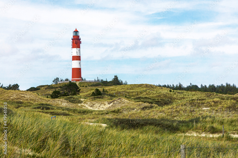 Tall red and white striped lighthouse building standing on small hill. Sandy coastal landscape with green vegetation and footpaths.