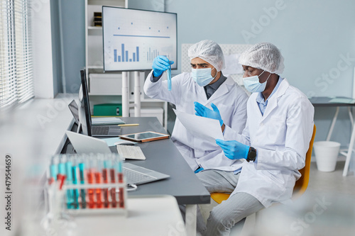 Two men in protective workwear sitting at the table and examining samples in test tubes during teamwork in the lab