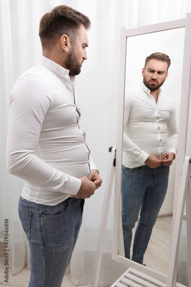 Upset man wearing tight shirt in front of mirror at home. Overweight problem Upset man wearing tight shirt in front of mirror at home. Overweight problem