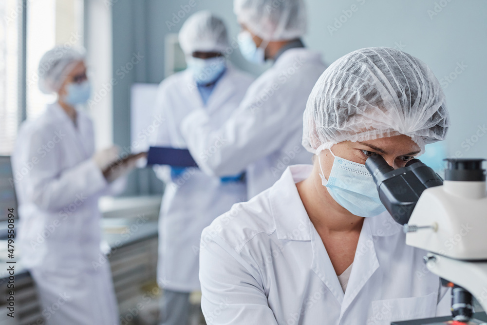 Young female scientist in protective mask sitting at the table and looking through the microscope, she working in the laboratory Young female scientist in protective mask sitting at the table and looking through the microscope, she working in the laboratory