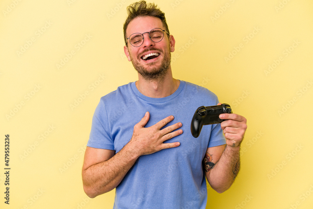 Young caucasian man holding a game controller isolated on yellow background laughs out loudly keeping hand on chest. Young caucasian man holding a game controller isolated on yellow background laughs out loudly keeping hand on chest.