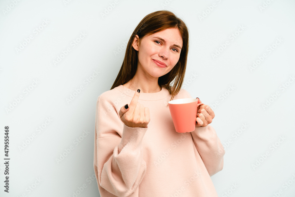 Young English woman holding a mug isolated on blue background pointing with finger at you as if inviting come closer. Young English woman holding a mug isolated on blue background pointing with finger at you as if inviting come closer.