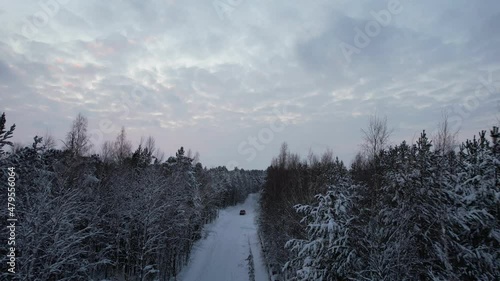 On a snowy road car rides in the middle of a winter frozen forest. The drone flies behind him at a low altitude. Firs and pines in the snow. Aerial view.