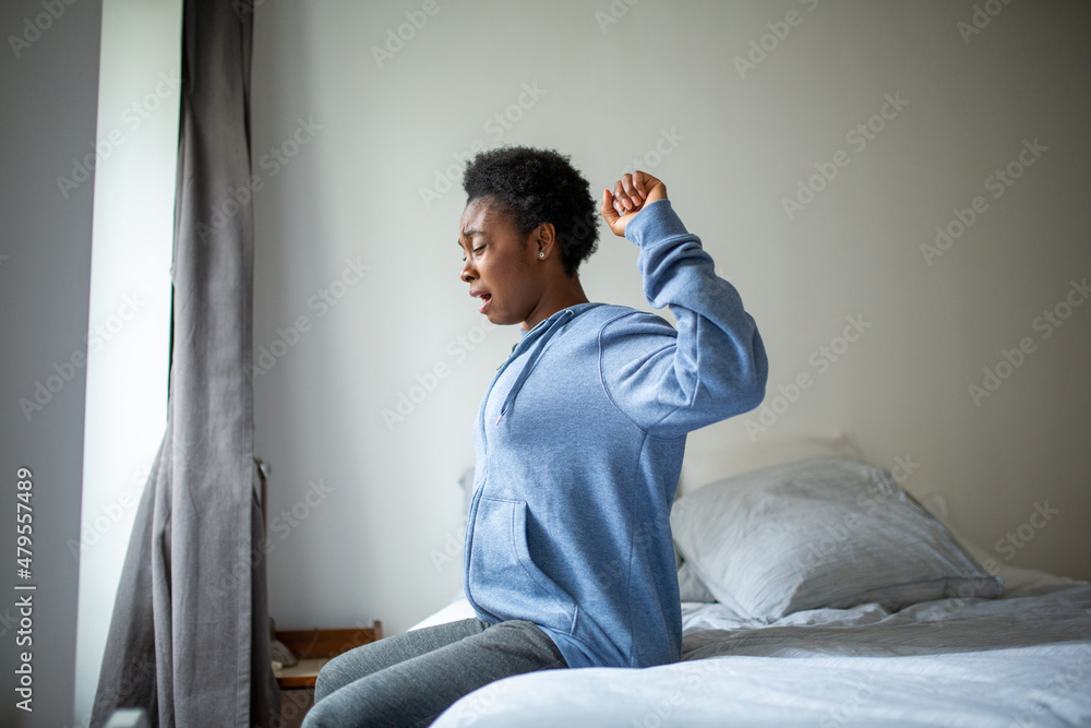 Side of tired young African American woman sitting on bed yawning with arms raised