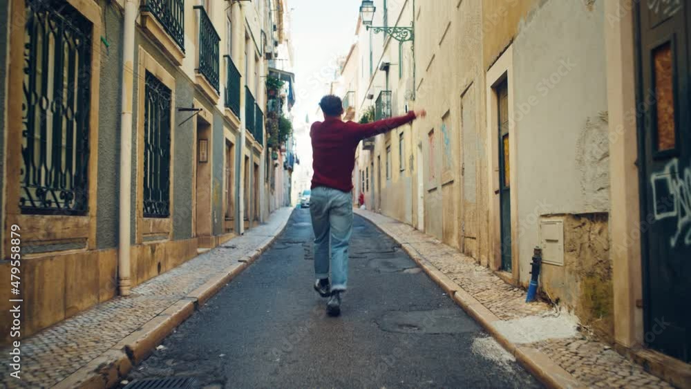 Cheerful and Happy Young Adult Man in Casual Clothes Actively Dancing while Walking on the Street of an Old Town in a City. Scene Shot in an Urban Quiet Narrow Town Street. Shot from Back. Cheerful and Happy Young Adult Man in Casual Clothes Actively Dancing while Walking on the Street of an Old Town in a City. Scene Shot in an Urban Quiet Narrow Town Street. Shot from Back.