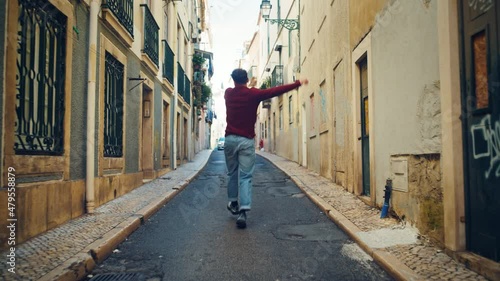 Cheerful and Happy Young Adult Man in Casual Clothes Actively Dancing while Walking on the Street of an Old Town in a City. Scene Shot in an Urban Quiet Narrow Town Street. Shot from Back.