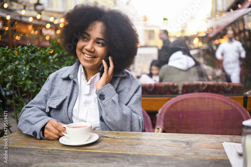 Fashion street style portrait of attractive young natural beauty African American woman with afro hair in tweed jacket speaks phone outdoors in sidewalk cafe. Happy lady with coffee cup in big city