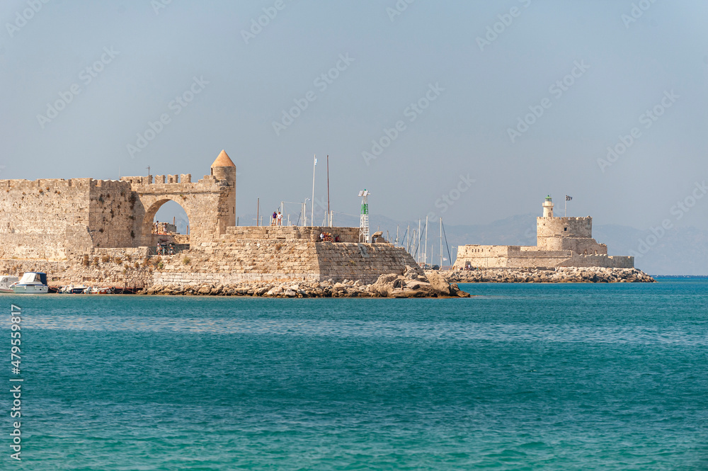 Die mittelalterliche Hafeneinfahrt von Rodos-Stadt auf der gleichnamigen griechischen Insel im Mittelmeer im Sommer bei Sonnenschein mit türkisfarbenen Meerwasser im Vordergrund