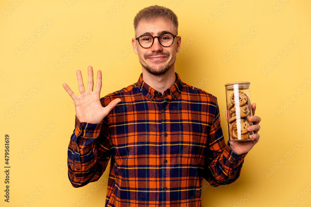 Young caucasian man holding cookies jar isolated on yellow background smiling cheerful showing number five with fingers. Young caucasian man holding cookies jar isolated on yellow background smiling cheerful showing number five with fingers.