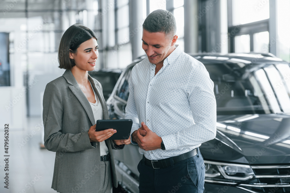 Standing near the car. Man in formal wear helping customer with choice of the automobile Standing near the car. Man in formal wear helping customer with choice of the automobile