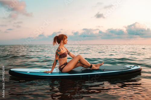 A tanned young woman with a tattoo poses sitting on a sup board swimming at water. Copy space. Sunset sky. The concept of sports recreation and surfing