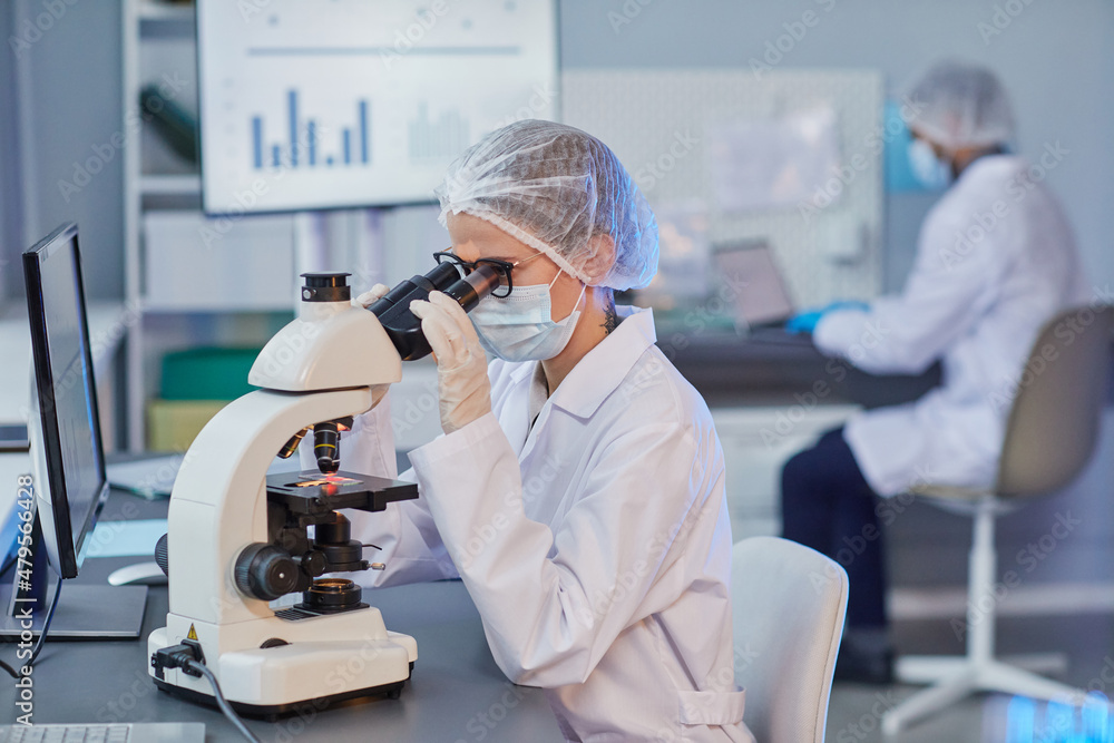 Young woman in lab coat looking through the microscope while sitting at her workplace, she examining sample working in the hospital lab Young woman in lab coat looking through the microscope while sitting at her workplace, she examining sample working in the hospital lab