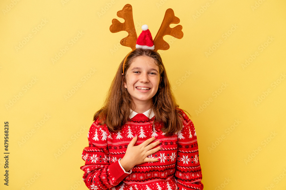Little caucasian girl wearing a Christmas reindeer hat isolated on yellow background laughs out loudly keeping hand on chest. Little caucasian girl wearing a Christmas reindeer hat isolated on yellow background laughs out loudly keeping hand on chest.