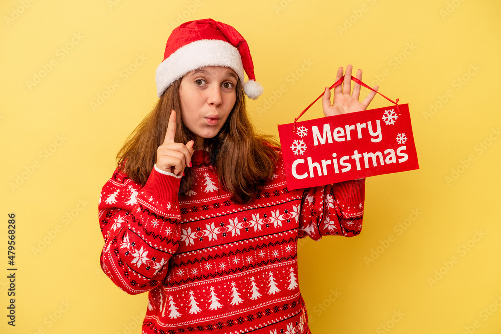 Little caucasian girl holding ‘’merry Christmas’’ placard isolated on yellow background having an idea, inspiration concept. Little caucasian girl holding ‘’merry Christmas’’ placard isolated on yellow background having an idea, inspiration concept.