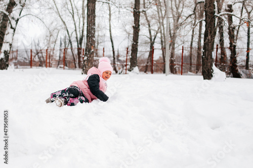 Winter portrait of female kid smiling and lying outside in snow in warm winter clothes with high trees around. Adorable background full of snow.