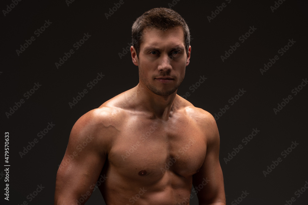 Fototapeta premium Waist up view of the portrait of shirtless bodybuilder looking at the camera while posing over the black wall. Stock photo