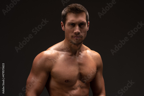 Waist up view of the portrait of shirtless bodybuilder looking at the camera while posing over the black wall. Stock photo