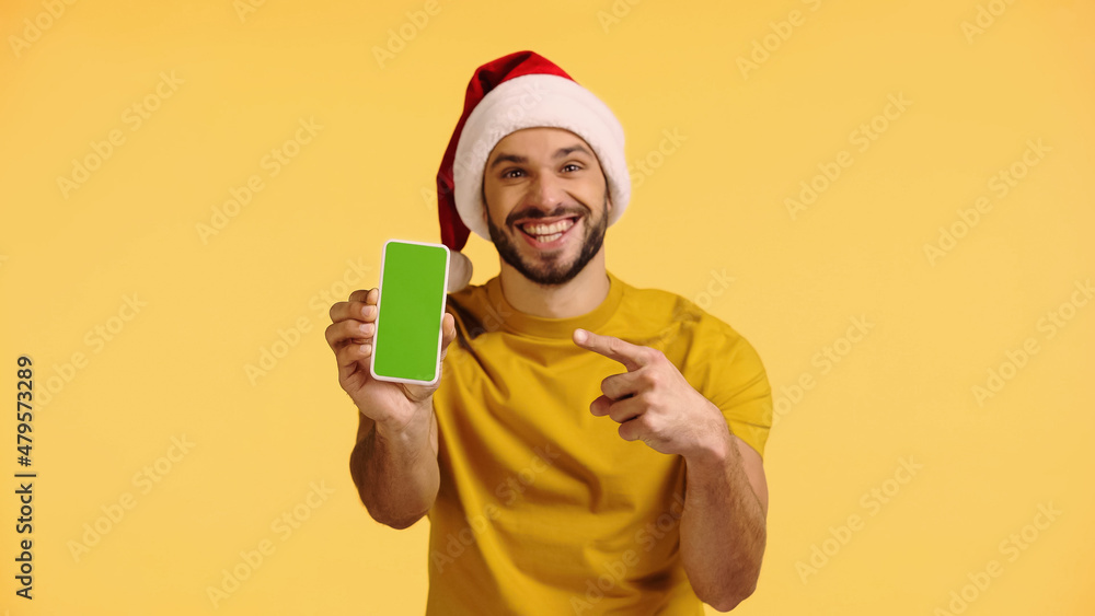 cheerful man in santa hat pointing at smartphone with green screen isolated on yellow cheerful man in santa hat pointing at smartphone with green screen isolated on yellow