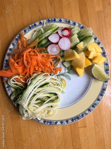 Close up of healthy vegan raw vegetable meal with carrot cucumber coriander radish mango lime onion and chilli sauce flat lay plate view on wood surface interior photo