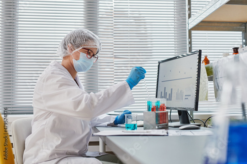 Young chemist in protective wear sitting at the table in front of computer monitor and working with test tubes with samples and analysis