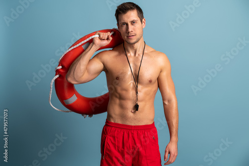Photos Positive man lifeguard with glad expression carries lifebuoy and looking at the camera with serious face isolated over blue background