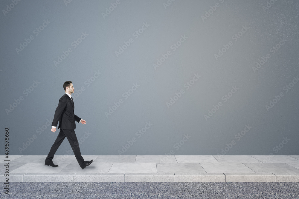 Side view of young business man walking in concrete interior with mock up place on wall. Success concept. Side view of young business man walking in concrete interior with mock up place on wall. Success concept.