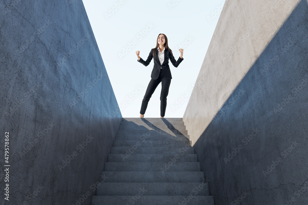 Happy european woman celebrating success on top of abstract concrete stairs with sunlight. Growth and leadership concept.