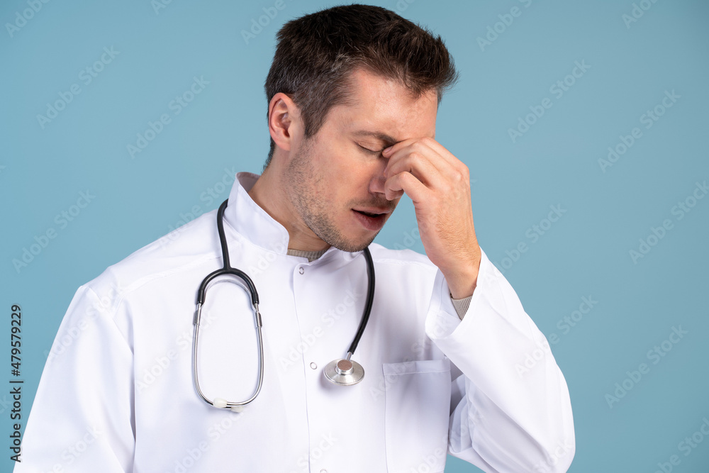 Portrait of upset depressed young man rubbing his nose and feeling headache after long hours of working. Indoor studio shot isolated on blue background Portrait of upset depressed young man rubbing his nose and feeling headache after long hours of working. Indoor studio shot isolated on blue background