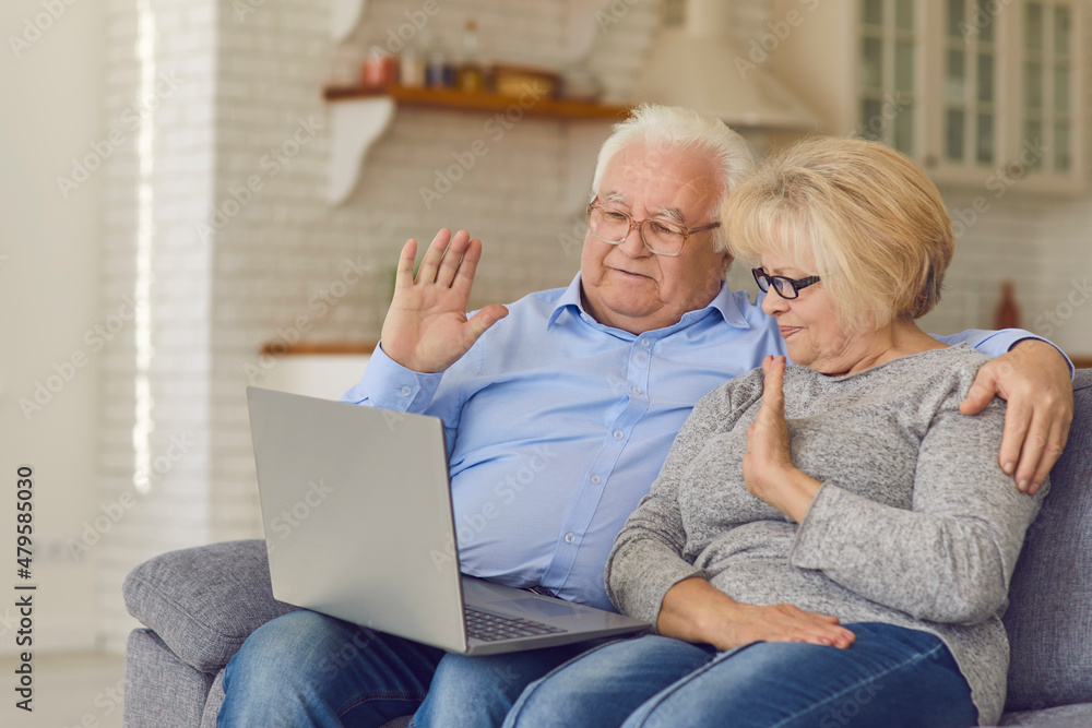 Happy grandparents staying home but keeping in touch with children and grandchildren. Smiling senior couple sitting on comfortable soft couch, video calling family and waving hands at laptop computer Happy grandparents staying home but keeping in touch with children and grandchildren. Smiling senior couple sitting on comfortable soft couch, video calling family and waving hands at laptop computer