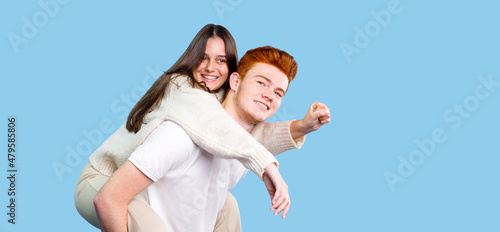 Two people brunette young woman and red hair young man making fun holding his girlfriend on the back over blue background, copy space.