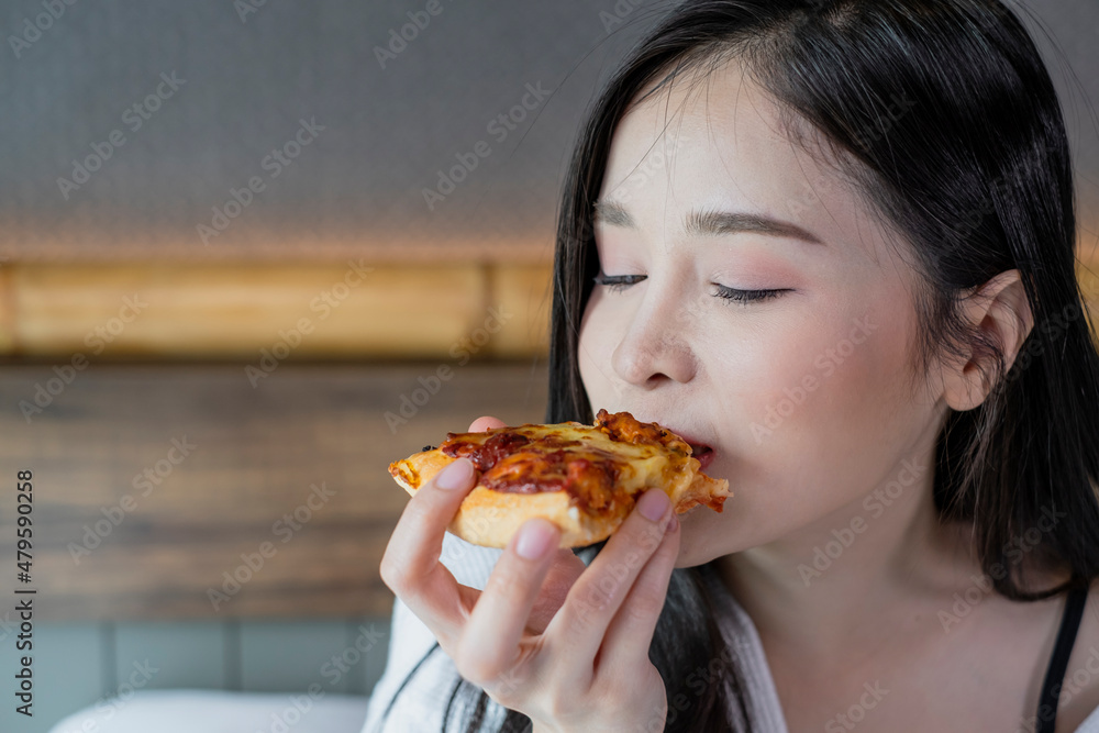 Happy young woman enjoy eating pizza on bed in hotel room. Beautiful female sitting in hotel bed eating pizza. Young woman hanging out eating pizza.