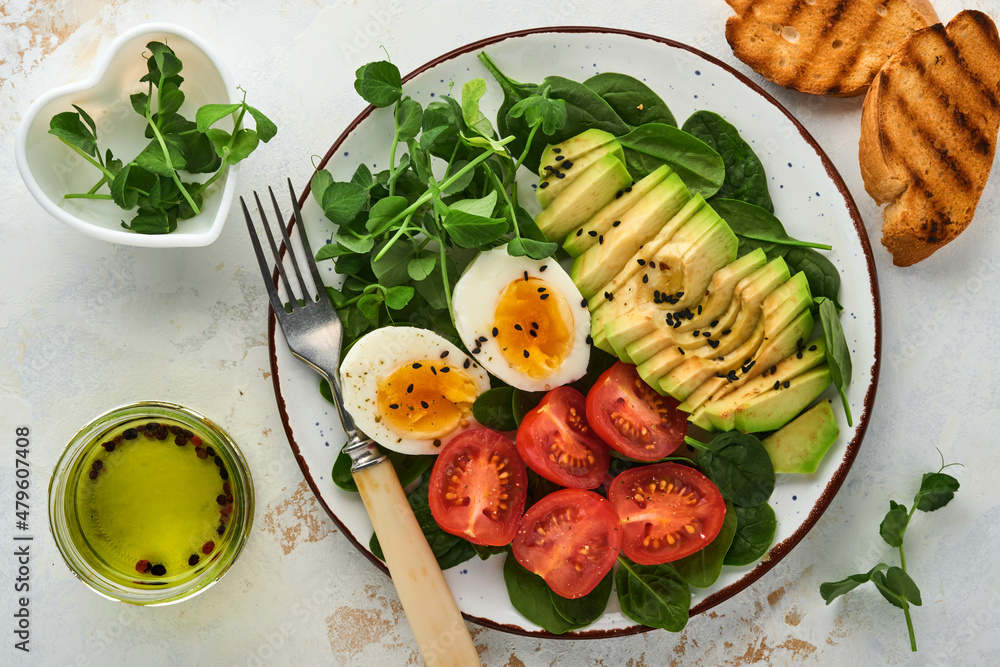 Avocado, cherry tomato, spinach and chicken egg, microgreens peas and black sesame seeds fresh salad in bowl on white stone table background. Healthy breakfast food concept. Top view. Avocado, cherry tomato, spinach and chicken egg, microgreens peas and black sesame seeds fresh salad in bowl on white stone table background. Healthy breakfast food concept. Top view.