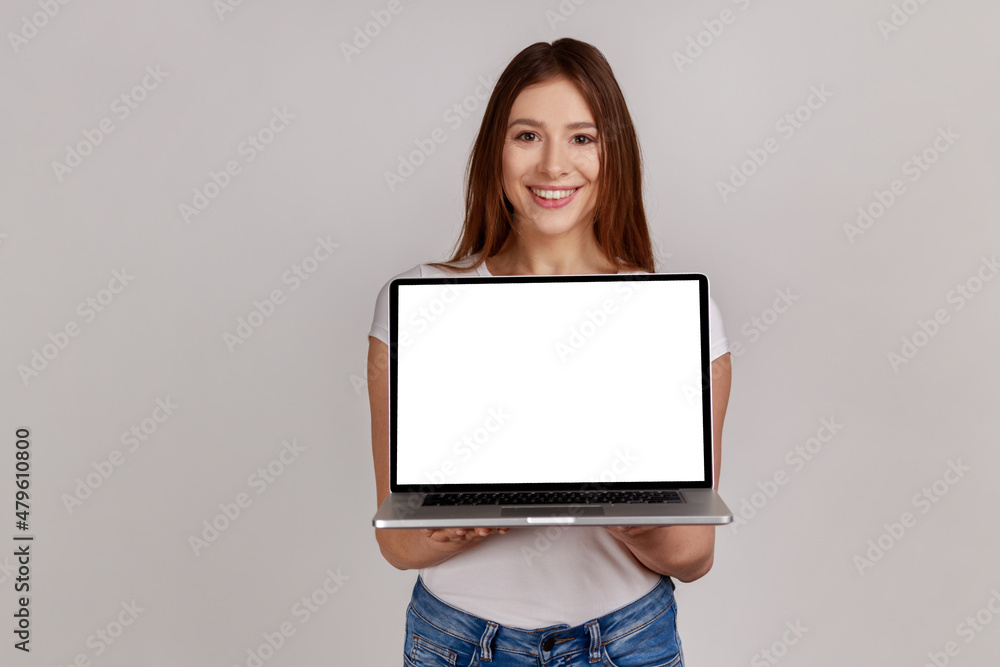Portrait of attractive smiling woman holds laptop with white display for internet advertising, online service, website promotion, wearing white T-shirt. Indoor studio shot isolated on gray background. Portrait of attractive smiling woman holds laptop with white display for internet advertising, online service, website promotion, wearing white T-shirt. Indoor studio shot isolated on gray background.