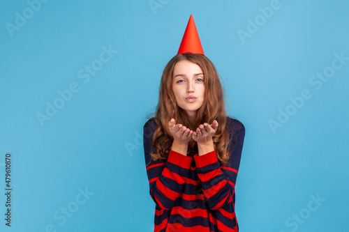 Pleased woman wearing striped casual style sweater and red cone sending air kisses, celebrating her birthday, expressing love to her guests. , Indoor studio shot isolated on blue background.