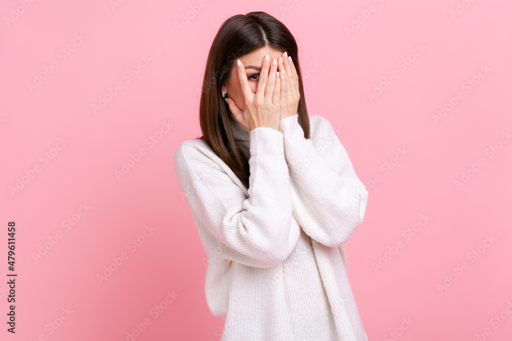 Curious woman looking through fingers, peeking with calm face, having suspicious, watching secrets, wearing white casual style sweater. Indoor studio shot isolated on pink background. Curious woman looking through fingers, peeking with calm face, having suspicious, watching secrets, wearing white casual style sweater. Indoor studio shot isolated on pink background.