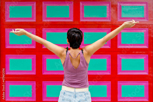 Cropped rear view of woman raising open arms against checkered wall decorated with red and green rectangles. Horizontal view of latin american woman isolated on colorful wall with summer clothes. photo